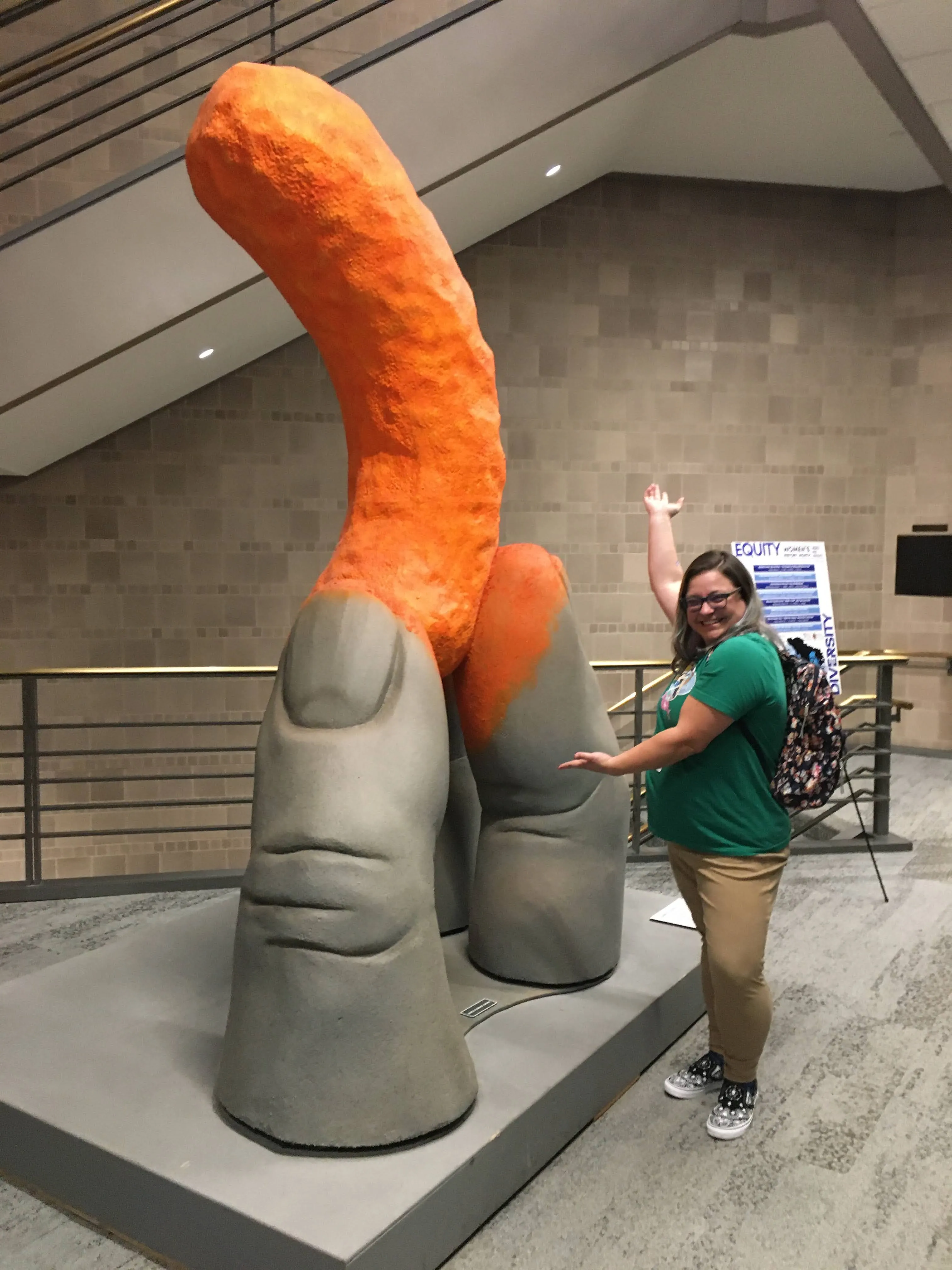 Linda poses with her arms outstretched towards a statue twice her size of a thumb, forefinger, and middle finger pointing towards the sky and holding a neon orange Cheetos puff, complete with cheese dust on the finger pads.