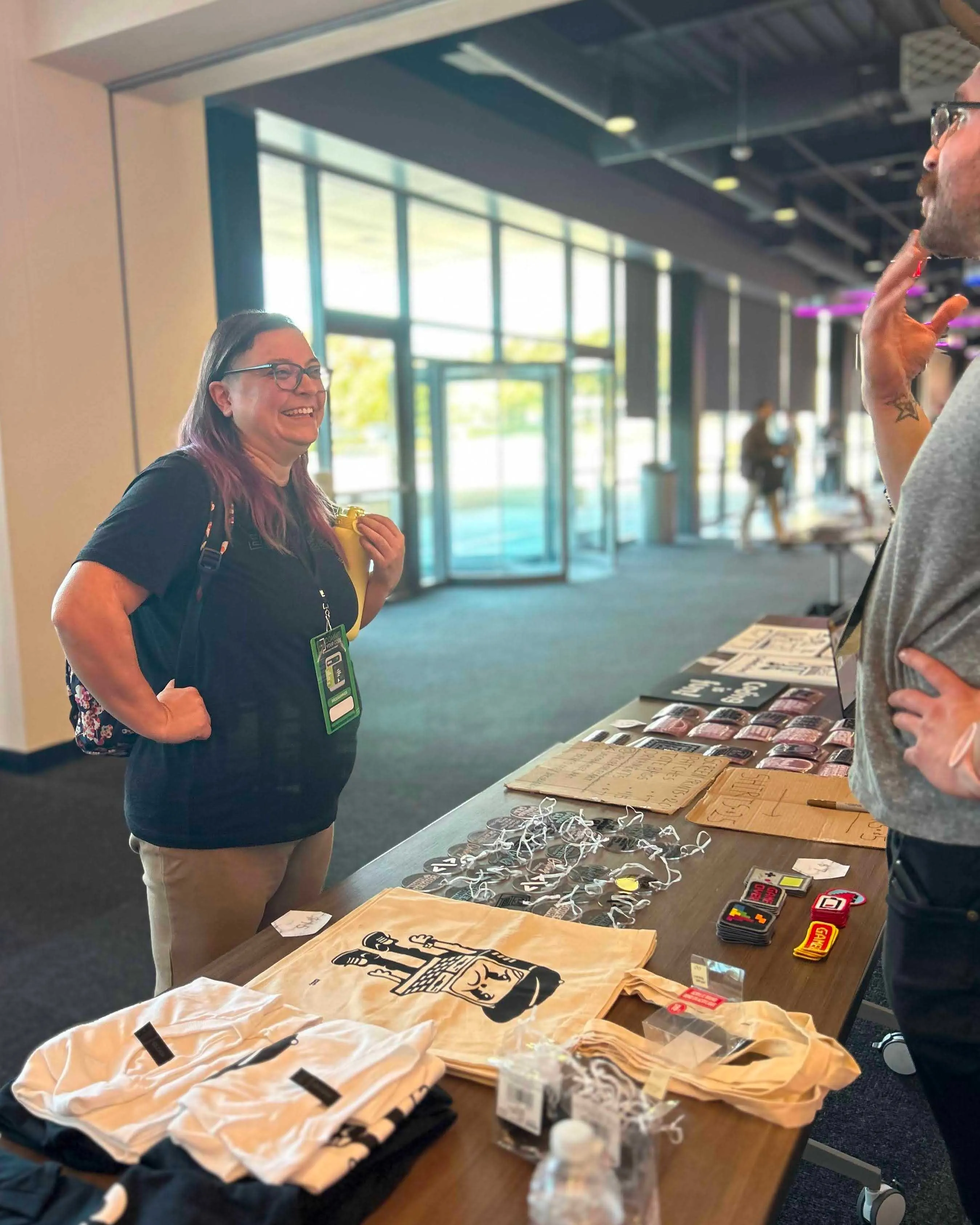 Linda and Jacob are mid conversation across a table filled with Jacob's artwork. There are bags with a sad computer on them, stickers galore, game patches, and more.