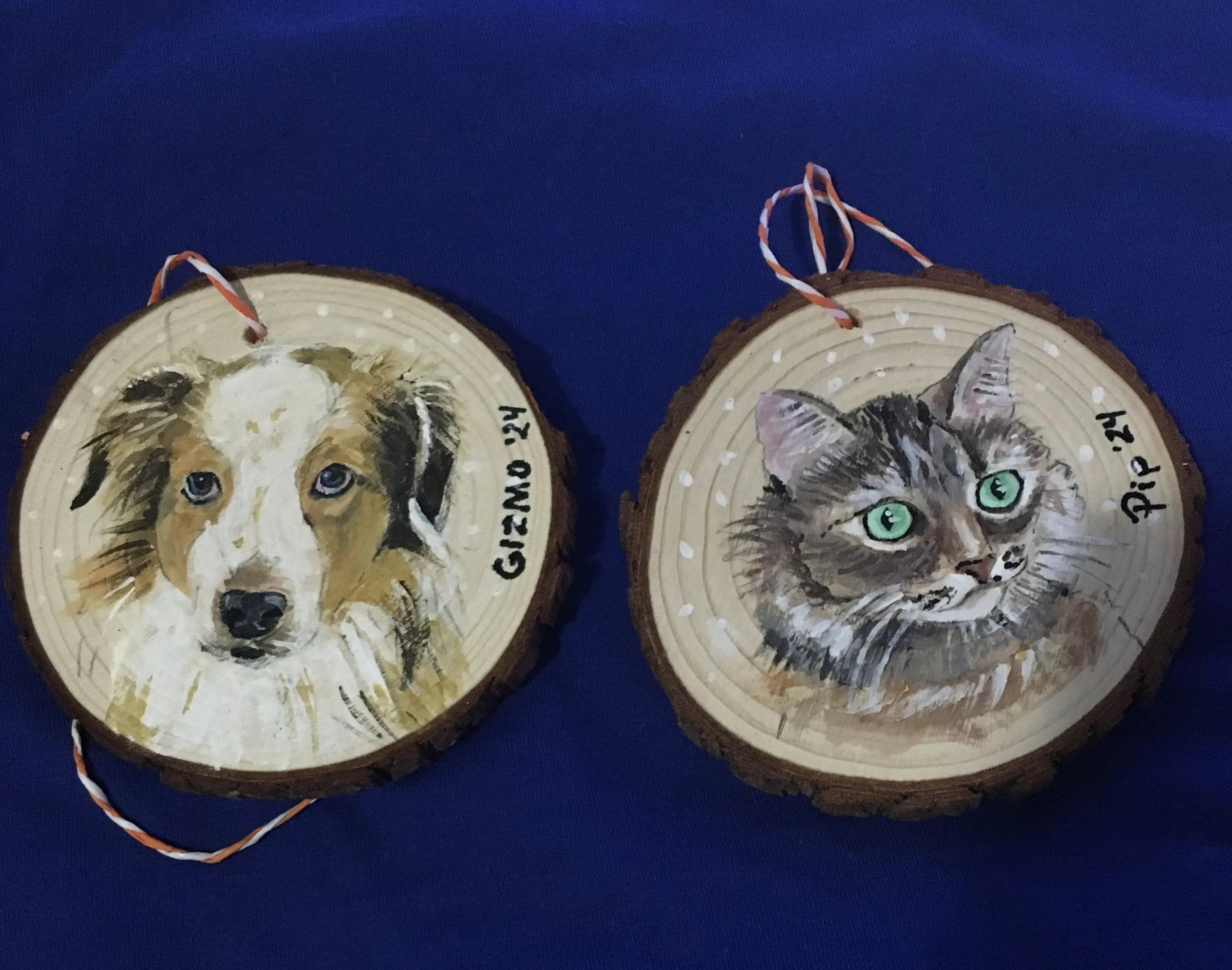 Two round wooden disks with a small hole in the top center with red and white string through it. The left disk has a painting of a tan and white english shepard. The right disk has a painting of a gray, tan, and white long haired cat. Their names are written along the right side of each disk, Gizmo on the left, Pip on the right.