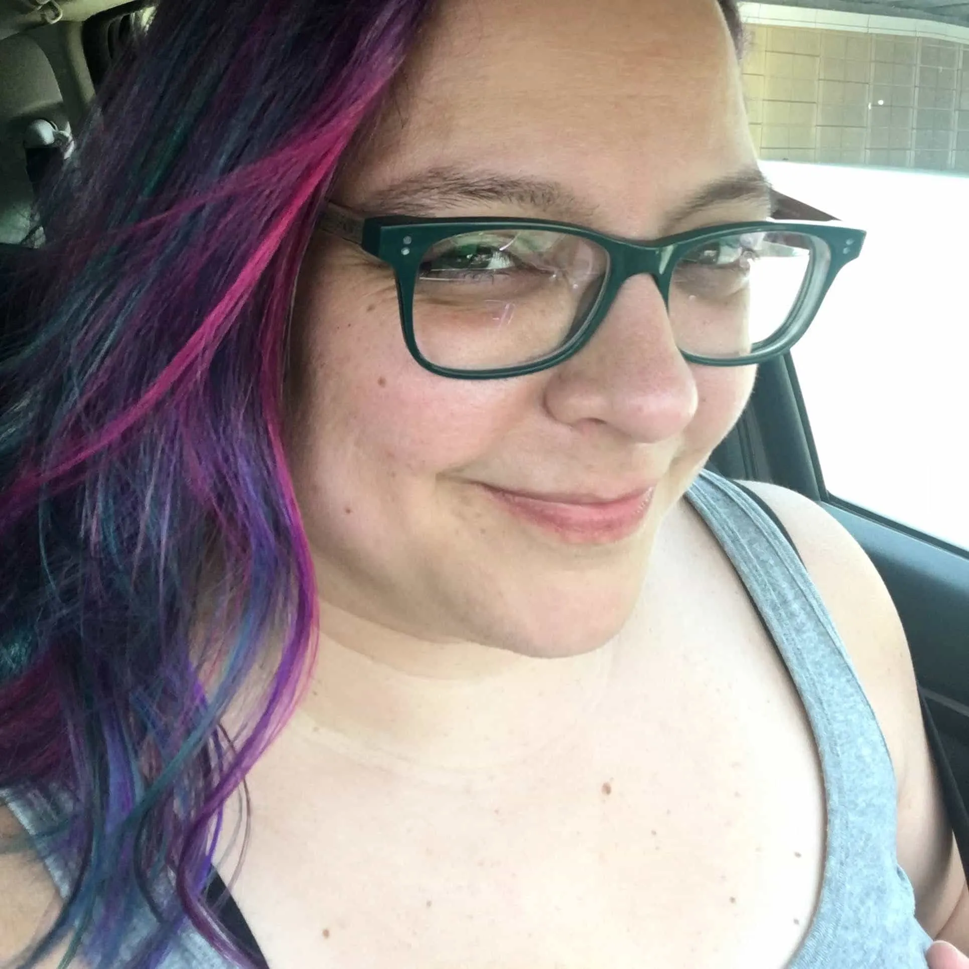 Woman with brown eyes, green glasses, and blue, purple, pink, and brown hair stares directly at you with a confident and friendly smirk.