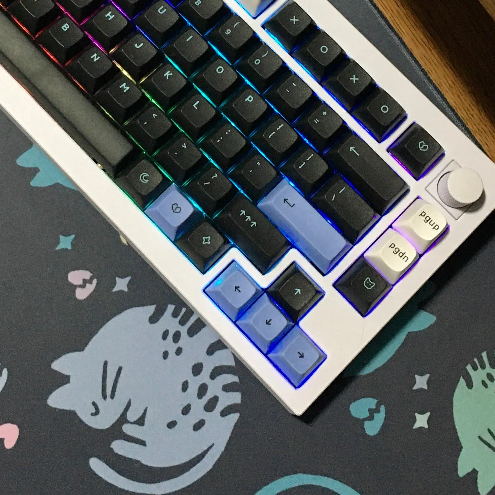 A white keyboard features a black, pastel blue, and white key cap set. The keyboard sits on top of a dark gray desk mat with curled sleeping cats in pastel blue and green and small broken hearts and stars in various pastel colors.