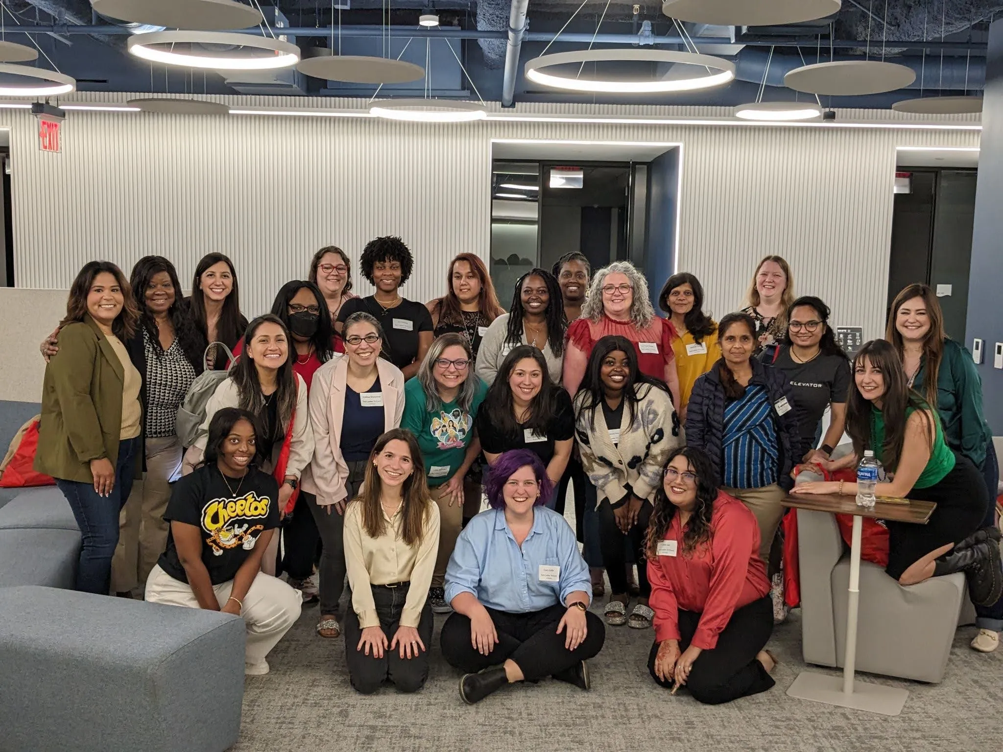 Four rows of ladies in tech stand together smiling for the camera in a bright room with white walls, gray carpet, and blue ceiling.