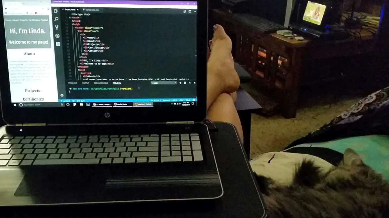 A soft cat sleeps on a multi-colored blanket on the right side of a laptop in a young woman's lap. The laptop has gray keys and shows a code editor and a portfolio page in progress on the screen.