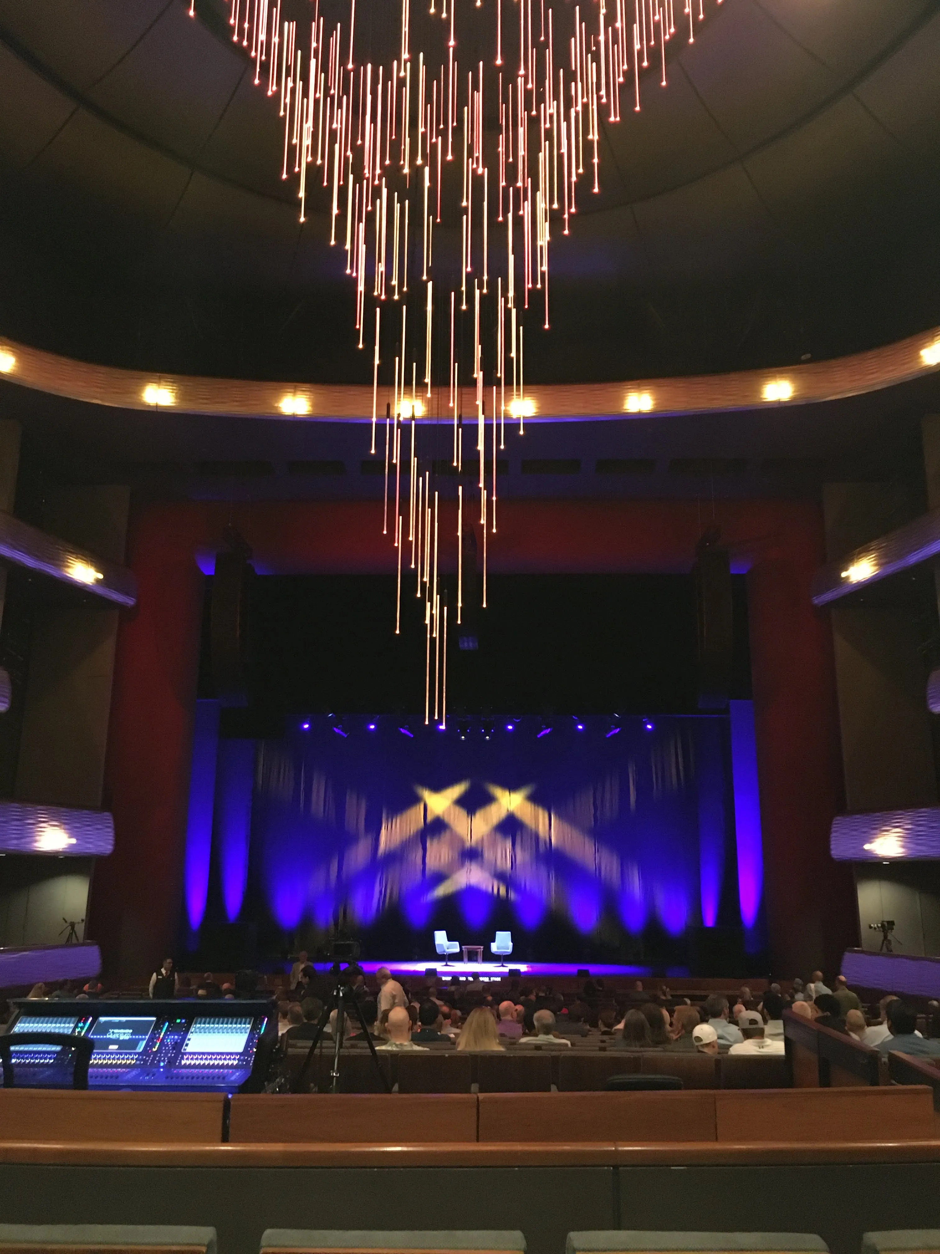 Center stage is a blue background dramatically lit, with two chairs and a table on the stage in front of it. Red walls and tan seating surrounds the stage. Hanging from the ceiling is a stunning chandelier made of long, thin rods of warm white light.